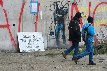 Des réfugiés passent devant un graffiti de l'artiste de rue Banksy représentant en migrant le fondateur d'Apple, Steve Jobs, à l'époque de l'évacuation complète du camp de la "Jungle" de Calais, à Calais, le 24 octobre 2016 - DENIS CHARLET [AFP]