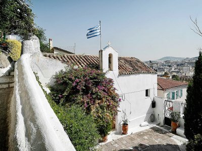 Un drapeau grec à côté de la croix d'une église à Anafiotika, un quartier situé dans la zone archéologique de l'Acropole, le 6 juillet 2018 à Athènes, en Grèce - LOUISA GOULIAMAKI [AFP]