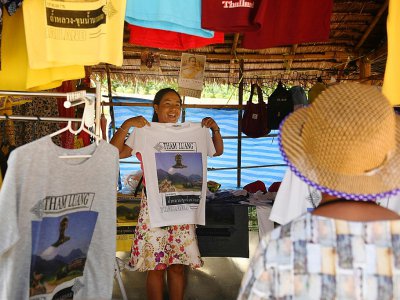 Une vendeuse montre à des touristes un "t-shirt souvenir" du sauvetage des adolescents en 2018 dans la grotte inondée de Tham Luang,  le 13 juin 2019 à Mae Sai, en Thaïlande - Lillian SUWANRUMPHA [AFP]