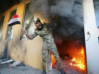Un membre des paramilitaires irakiens pro-iraniens du Hachd al-Chaabi  arbore un drapeau irakien en sortant d'une salle en flammes après avoir forcé le mur d'enceinte de l'ambassade des Etats-Unis à Bagdad, le 31 décembre 2019 - AHMAD AL-RUBAYE [AFP]