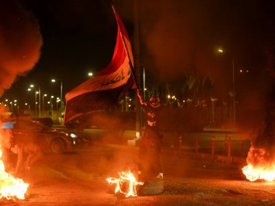 Un manifestant brandit le drapeau irakien devant un pneu en flammes sur une artère de la ville de Najaf, au sud de Bagdad, le 1er février 2020 au soir - Haidar HAMDANI [AFP]