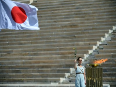 La flamme olympique devant le drapeau du Japon, pendant la cérémon ie de passation de la flamme entre la Grèce et le Japon dans un stade panathénaïque vide, le 19 mars 2020 à Athènes - ARIS MESSINIS [AFP]
