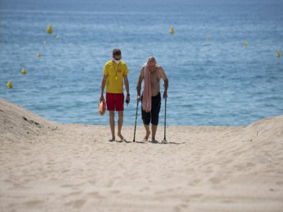 Un secouriste, portant un masque de protection, accompagne un homme âgé sur la plage de Lloret de Mar, le 22 juin 2020 en Espagne - Josep LAGO [AFP]