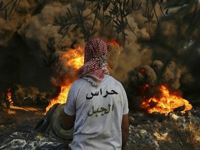 Un Palestinien portant un tee-shirt barré sur le dos de l'inscription "Gardiens de la montagne" devant des pneus brûlés dans le village de Beita face à la colonie d'Eviatar en Cisjordanie, le 23 juin 2021 - ABBAS MOMANI [AFP]