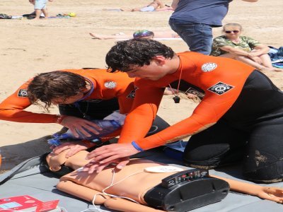 Une fois la victime extraite de l'eau, elle est remplacée par un mannequin pour vérifier l'apprentissage délicat du massage cardiaque. Une autre membre de l'équipe a préparé le défibrillateur sur la plage.